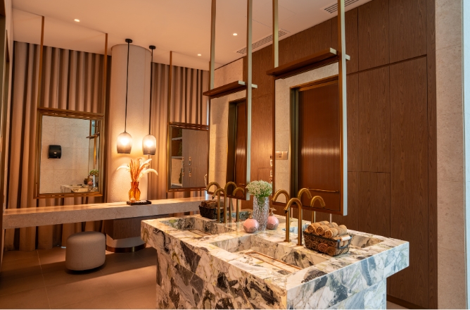 http://A%20luxurious%20spa%20vanity%20room%20featuring%20dual%20integrated%20green-and-white%20marble%20block%20sinks%20with%20elegant%20brass%20faucets,%20hanging%20bronze-framed%20mirrors,%20and%20a%20soft-lit%20powder%20area.