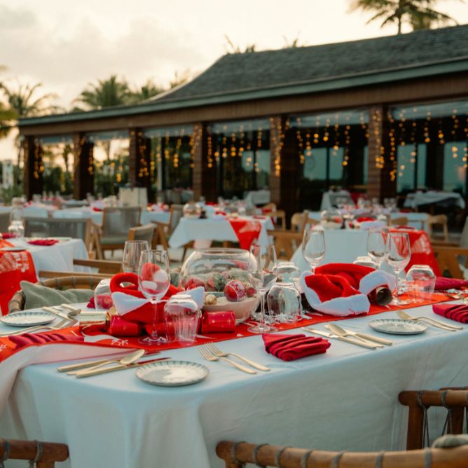 Outdoor resort restaurant tables set for a Christmas dinner with red runners, Santa hats, and festive centerpieces near the beach.