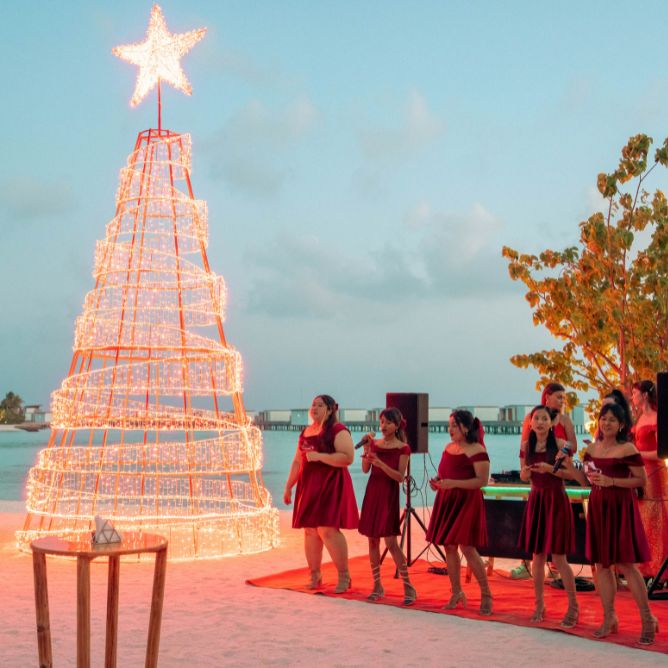 Group of women in red dresses singing next to a large spiral light Christmas tree on a sandy beach during a holiday celebration.