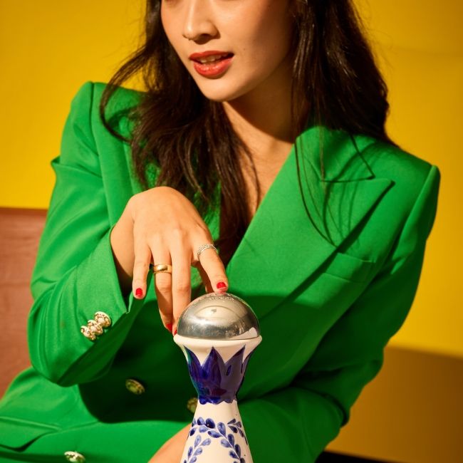 A woman in a green suit holds a decorative vase, showcasing her elegant style and the vibrant colors of the arrangement.